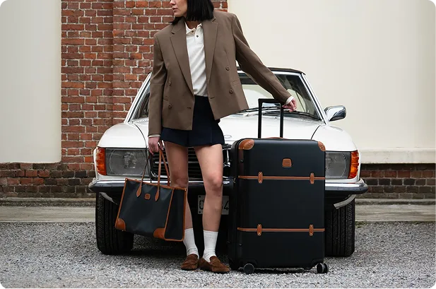 Woman in a tan blazer holding a designer tote bag next to a black wheeled suitcase and a vintage car